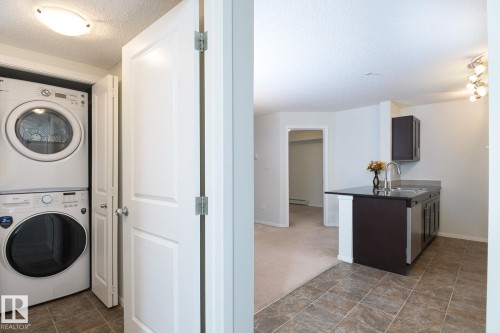 Compact laundry area featuring a stacked washer and dryer unit, complemented by a kitchen space with dark cabinetry, stainless steel sink, and a black countertop - 111 25 Element Dr, St. Albert, AB - Indoor Photo Showing Laundry Room