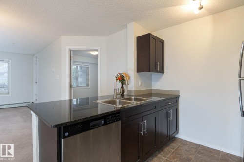 Kitchen featuring dark wood-finish cabinetry, granite-style countertops, double stainless steel sink, and stainless steel dishwasher - 111 25 Element Dr, St. Albert, AB - Indoor Photo Showing Kitchen With Double Sink