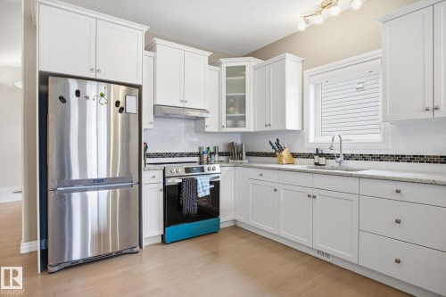 Kitchen featuring white cabinetry with crown molding, stainless steel appliances, a white subway tile backsplash with a mosaic accent, light-toned countertops, and wood-finish flooring - 28 Hewitt Circle, Spruce Grove, AB - Indoor Photo Showing Kitchen With Stainless Steel Kitchen