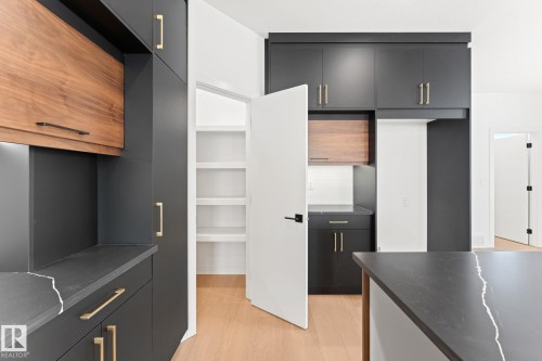 Modern kitchen featuring dark cabinetry with wood accents, gold-toned hardware, light hardwood flooring, and a pantry with shelving - 94 Blackbird Bend, Fort Saskatchewan, AB - Indoor