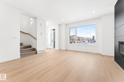 The property features an open-concept living space with light-toned hardwood flooring, a large window providing ample natural light, and a modern fireplace with a dark surround - 94 Blackbird Bend, Fort Saskatchewan, AB - Indoor