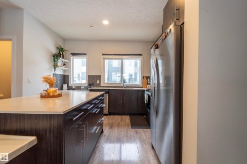 Kitchen featuring a large central island with a light-toned countertop, dark wood-finish cabinetry with silver hardware, wood-finish flooring, a stainless steel refrigerator, and a dark tile backsplash - 117 804 Welsh Drive, Edmonton, AB - Indoor Photo Showing Kitchen