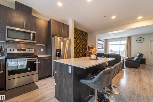 Modern kitchen featuring dark wood-finish cabinetry, stainless steel appliances, a white countertop island with seating, and light wood-finish flooring - 117 804 Welsh Drive, Edmonton, AB - Indoor Photo Showing Kitchen With Upgraded Kitchen