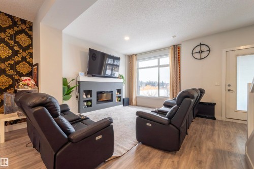 Living area featuring wood-finish flooring, a contemporary fireplace with integrated shelving, and a large window with roller shades - 117 804 Welsh Drive, Edmonton, AB - Indoor Photo Showing Living Room With Fireplace