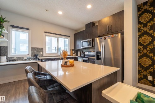 Modern kitchen featuring a large island with a light-toned countertop, stainless steel appliances, dark wood-finish cabinetry, a herringbone tile backsplash, and recessed lighting - 117 804 Welsh Drive, Edmonton, AB - Indoor Photo Showing Kitchen