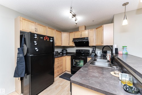 L-shaped kitchen featuring wood-finish cabinetry, dark countertops, and black appliances - 219 646 Mcallister Loop, Edmonton, AB - Indoor Photo Showing Kitchen With Double Sink
