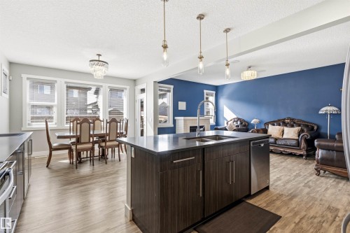 Open-concept living area featuring wood-finish flooring throughout, a central kitchen island with an undermount double sink, a dining area with a multi-pane window, and a spacious living room with a feature wall and fireplace - 9812 224 Street, Edmonton, AB - Indoor