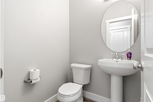Powder room featuring a pedestal sink with a chrome faucet, a round wall-mounted mirror, and wood-finish flooring - 9812 224 Street, Edmonton, AB - Indoor Photo Showing Bathroom