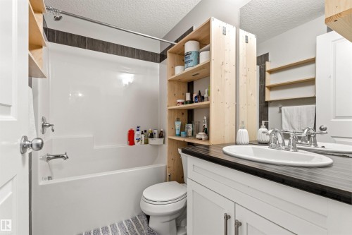 Bathroom featuring a white vanity with a dark countertop, an oval undermount sink, a white tub/shower combination with dark accent tiling, and natural wood shelving - 9812 224 Street, Edmonton, AB - Indoor Photo Showing Bathroom