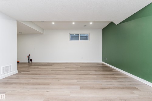 Spacious room featuring light wood-finish flooring, recessed lighting, a window with blinds, a contrasting green accent wall, and white baseboards - 16420 15 Avenue, Edmonton, AB - Indoor