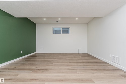Spacious room featuring wood-finish flooring, recessed lighting, a window with horizontal blinds, and a contrasting green accent wall - 16420 15 Avenue, Edmonton, AB - Indoor Photo Showing Other Room