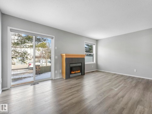 The living area features light grey walls, grey wood-style flooring, a modern fireplace with a light wood mantel, and large sliding glass doors providing natural light - 175 150 Edwards Drive, Edmonton, AB - Indoor Photo Showing Living Room With Fireplace