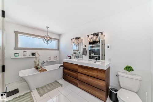 Bathroom featuring a freestanding soaking tub, wood-finish double vanity with dual sinks, overhead geometric pendant light fixture, and white tile flooring - 5263 47 Ave, Calmar, AB - Indoor Photo Showing Bathroom