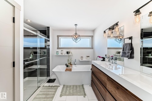 Contemporary bathroom featuring a freestanding soaking tub, a frameless glass-enclosed shower with dark tile surround, a large vanity with wood-finish cabinetry and white countertop, and modern industrial-style lighting fixtures - 5263 47 Ave, Calmar, AB - Indoor Photo Showing Bathroom