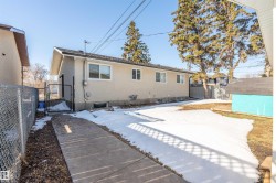 Rear exterior featuring a neutral stucco finish, multiple windows, a chain-link fence, a concrete walkway, and mature trees in the background - 