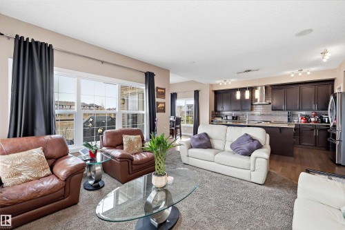 Open-concept living space featuring a carpeted living area, a kitchen with dark cabinetry and stainless steel appliances, a tile backsplash, wood-finish flooring, and a large window overlooking the neighborhood - 1586 Chapman Way, Edmonton, AB - Indoor Photo Showing Living Room