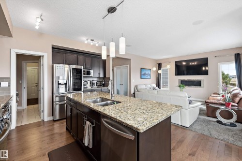 Open-concept kitchen featuring an oversized island with an integrated sink, granite countertops, dark wood cabinetry, and stainless steel appliances - 1586 Chapman Way, Edmonton, AB - Indoor Photo Showing Kitchen With Fireplace With Double Sink With Upgraded Kitchen