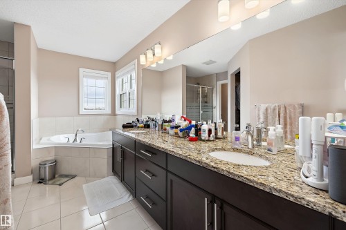 Bathroom featuring a dual vanity with granite-style countertops and wood-finish cabinetry, a large built-in mirror with integrated lighting, a corner soaking tub with tile surround, and a glass-enclosed shower - 1586 Chapman Way, Edmonton, AB - Indoor Photo Showing Bathroom