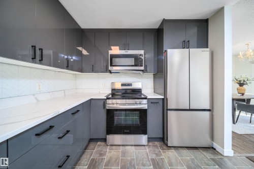 Modern kitchen featuring grey high-gloss cabinetry, stainless steel appliances, white patterned tile backsplash, white countertops, and wood-look tile flooring - 5303 21 Avenue, Edmonton, AB - Indoor Photo Showing Kitchen