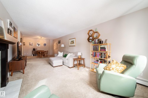 Carpeted living area featuring a dark wood mantle, a baseboard heating unit, and neutral wall tones - 220 14819 51 Avenue, Edmonton, AB - Indoor Photo Showing Other Room