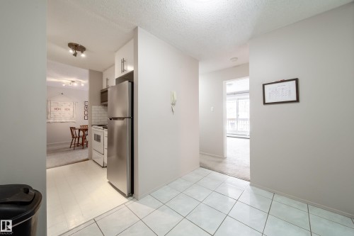 Crisp white tile flooring defines this entry area, leading to a compact kitchen featuring stainless steel appliances and white cabinetry - 220 14819 51 Avenue, Edmonton, AB - Indoor