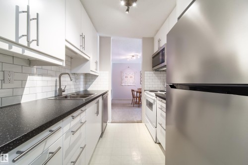 Galley kitchen featuring white cabinetry with brushed nickel hardware, dark speckled countertops, subway tile backsplash, and stainless steel appliances - 220 14819 51 Avenue, Edmonton, AB - Indoor Photo Showing Kitchen With Double Sink