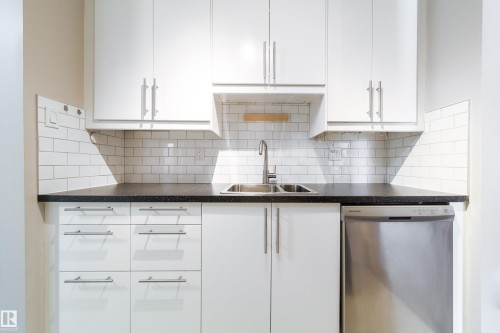 Contemporary kitchen with white cabinetry, a double basin stainless steel sink, a stainless steel dishwasher, dark speckled countertops, and white subway tile backsplash - 220 14819 51 Avenue, Edmonton, AB - Indoor Photo Showing Kitchen With Double Sink