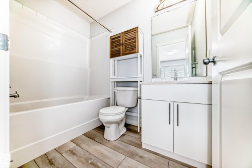 Bathroom featuring wood-finish flooring, a white integrated tub and shower unit, a white vanity with a light-toned countertop, a rectangular mirror, and a wall-mounted light fixture - 2505 14 Avenue, Edmonton, AB - Indoor Photo Showing Bathroom