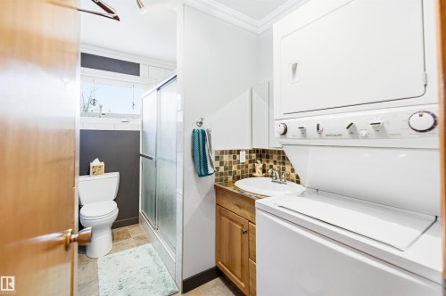 Compact bathroom featuring a stacked washer and dryer, wood-finish vanity with an undermount sink, mosaic tile backsplash, glass-enclosed shower, and a white toilet - 3920 35A Avenue, Red Deer, AB - Indoor Photo Showing Laundry Room