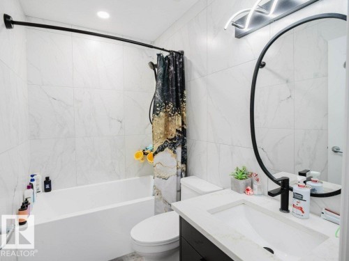 Contemporary bathroom featuring white tile walls, a built-in bathtub, a vanity with an integrated sink and white countertop, a circular black-framed mirror, and modern overhead lighting - 5701 Rue Eaglemont, Beaumont, AB - Indoor Photo Showing Bathroom