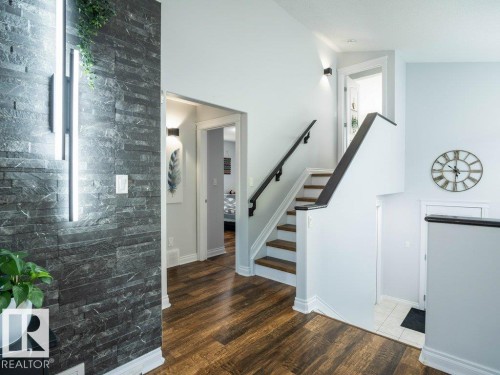 Interior entryway featuring a dark stacked stone accent wall with integrated vertical lighting, wood-finish flooring, a contemporary staircase with dark wood handrails, light-colored wall paint, and white trim throughout - 5701 Rue Eaglemont, Beaumont, AB - Indoor Photo Showing Other Room