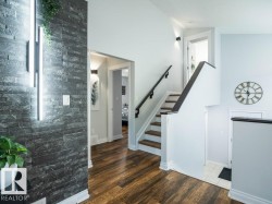 Interior entryway featuring a dark stacked stone accent wall with integrated vertical lighting, wood-finish flooring, a contemporary staircase with dark wood handrails, light-colored wall paint, and white trim throughout - 