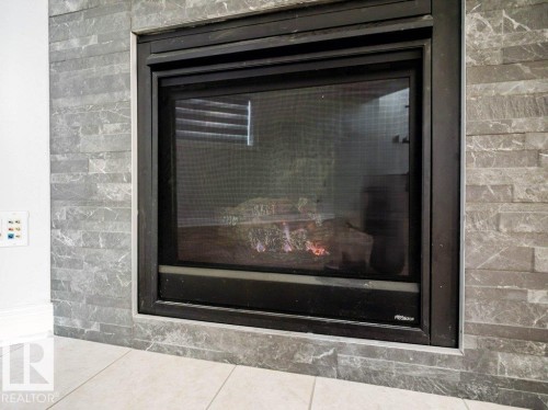 Modern fireplace featuring a black metal frame, gray stacked stone surround, and light tile flooring - 5701 Rue Eaglemont, Beaumont, AB - Indoor Photo Showing Other Room