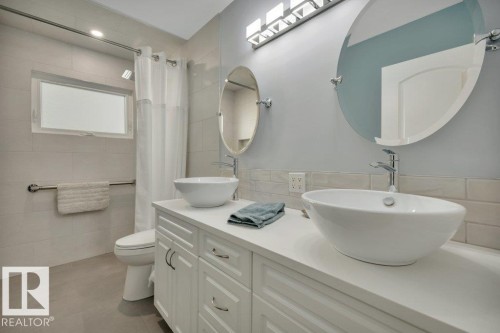 Dual vessel sinks with chrome faucets, white vanity cabinetry with chrome hardware, oval frameless mirrors, and a tiled shower surround with a window - 10633 St Gabriel School Road, Edmonton, AB - Indoor Photo Showing Bathroom