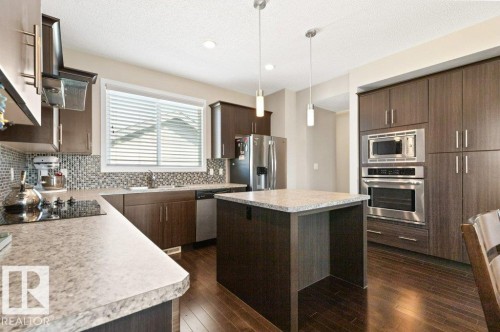 Modern kitchen featuring dark wood-finish cabinetry, stainless steel appliances, a center island with a light-toned countertop, a mosaic tile backsplash, and wood-finish flooring - 575 Allard Boulevard, Edmonton, AB - Indoor Photo Showing Kitchen With Upgraded Kitchen