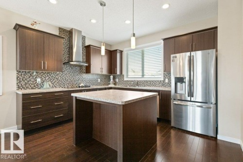 Kitchen featuring dark wood-finish cabinetry, a mosaic tile backsplash, stainless steel appliances, and a central island with a light-toned countertop - 575 Allard Boulevard, Edmonton, AB - Indoor Photo Showing Kitchen With Upgraded Kitchen