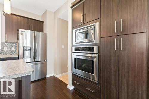 Kitchen featuring dark wood-finish cabinetry with brushed nickel hardware, stainless steel appliances including a built-in microwave and oven, a three-door refrigerator with dispenser, and a mosaic tile backsplash - 575 Allard Boulevard, Edmonton, AB - Indoor Photo Showing Kitchen