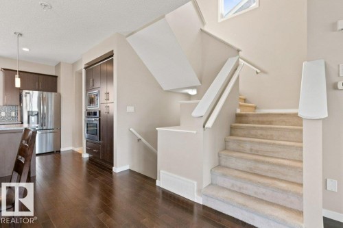 Open-concept living area featuring a carpeted staircase with white railings, rich wood-finish flooring, and a kitchen with dark cabinetry, stainless steel appliances, and a tiled backsplash - 575 Allard Boulevard, Edmonton, AB - Indoor Photo Showing Kitchen