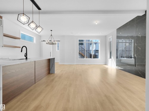 Open-concept living space featuring light wood-finish flooring, a kitchen island with a waterfall countertop and built-in sink, and a dark marble-look accent wall - 6722 Crawford Way, Edmonton, AB - Indoor