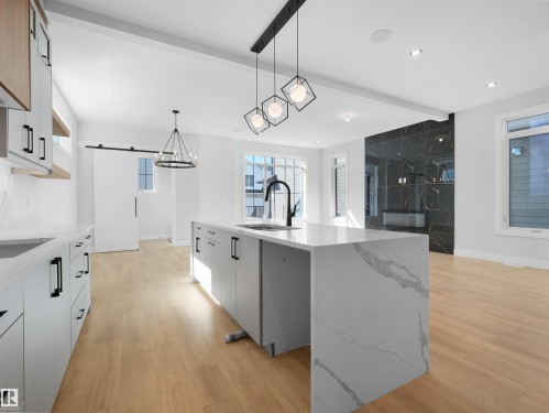 Open-concept kitchen and living area featuring a waterfall island with a sink and matte black faucet, light wood-finish flooring, white cabinetry with black hardware, and a black marble-style fireplace surround - 6722 Crawford Way, Edmonton, AB - Indoor Photo Showing Kitchen With Upgraded Kitchen