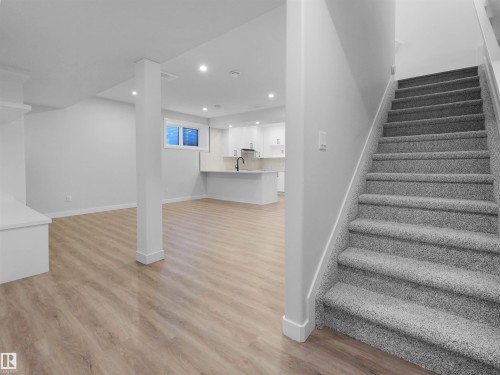 Gray carpeted staircase leading to a spacious area featuring wood-finish flooring, recessed lighting, and a white kitchen with a peninsula, light countertops, and a black faucet - 6722 Crawford Way, Edmonton, AB - Indoor Photo Showing Other Room