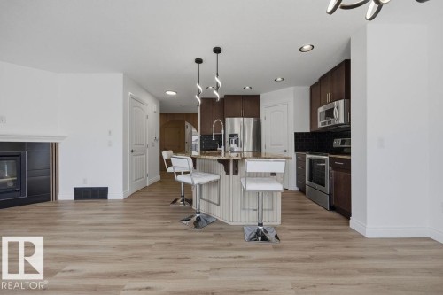 Open-concept kitchen and living area featuring wood-finish flooring, a central island with pendant lighting, stainless steel appliances, dark cabinetry, and a fireplace with a black surround - Edmonton, AB - Indoor Photo Showing Kitchen With Stainless Steel Kitchen With Upgraded Kitchen