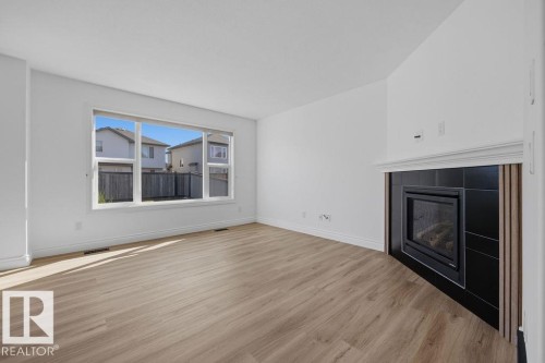 Spacious living area featuring light wood-finish flooring, a large window, and a corner-set fireplace with a black tile surround and white mantel - Edmonton, AB - Indoor Photo Showing Living Room With Fireplace