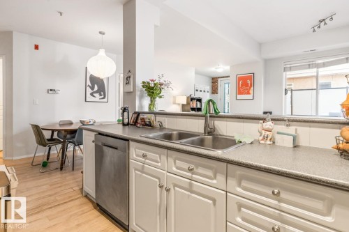Kitchen featuring a double basin stainless steel sink, a stainless steel dishwasher, white cabinetry with silver hardware, a tiled backsplash, and light-colored countertops - 109 10728 82 Avenue, Edmonton, AB - Indoor Photo Showing Kitchen With Double Sink