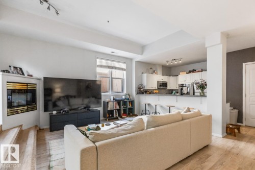 Open-concept living area featuring wood-finish flooring, a built-in fireplace, track lighting, a kitchen bar, and white cabinetry with stainless steel appliances - 109 10728 82 Avenue, Edmonton, AB - Indoor Photo Showing Living Room