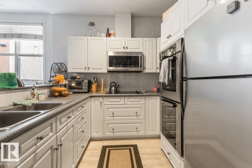 Bright kitchen featuring white shaker cabinetry, stainless steel appliances, a dual basin sink, light-toned flooring, and a window with blinds - 109 10728 82 Avenue, Edmonton, AB - Indoor Photo Showing Kitchen With Stainless Steel Kitchen With Double Sink