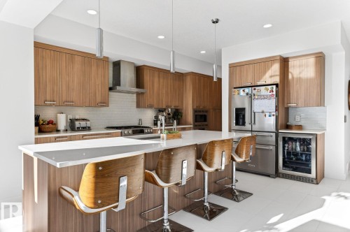 Modern kitchen featuring an expansive island with a white countertop, wood-finish cabinetry, stainless steel appliances, a tile backsplash, and recessed and pendant lighting - 793 Windermere Wynd, Edmonton, AB - Indoor Photo Showing Kitchen With Upgraded Kitchen