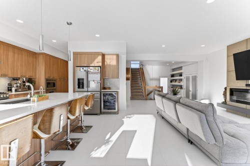 Open-concept living area featuring a kitchen island with seating, wood-finish cabinetry, stainless steel appliances, a modern fireplace, and a light wood-finish staircase - 793 Windermere Wynd, Edmonton, AB - Indoor With Fireplace