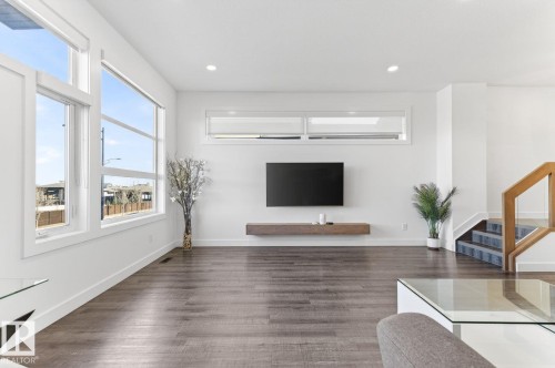 Spacious living area featuring expansive windows, wood-finish flooring, a wall-mounted television, a floating wood-finish shelf, and a staircase with wood railings and dark-toned treads - 793 Windermere Wynd, Edmonton, AB - Indoor