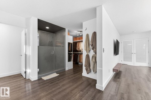 Modern open-concept interior featuring wood-finish flooring, a frameless glass shower enclosure with dark tile, a walk-in closet with built-in shelving, a wall-mounted display shelf, and frosted double French doors - 793 Windermere Wynd, Edmonton, AB - Indoor Photo Showing Other Room